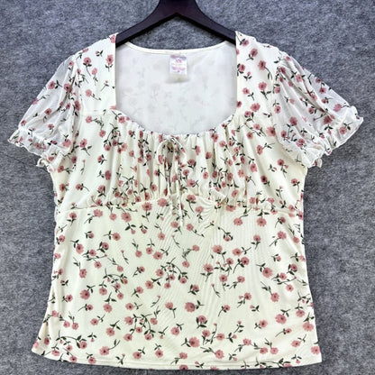 A cream-colored short-sleeve blouse with a pink and green floral print, featuring a square neckline with ruffles and a smocked bust, displayed on a black hanger against a gray textured background.