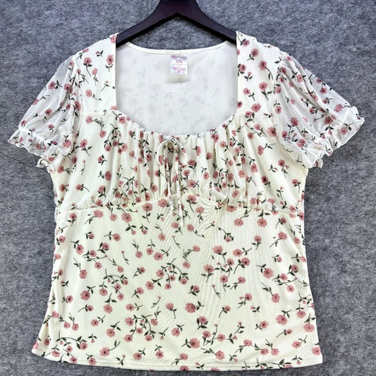 A cream-colored short-sleeve blouse with a pink and green floral print, featuring a square neckline with ruffles and a smocked bust, displayed on a black hanger against a gray textured background.