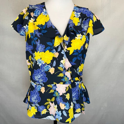 A women's short-sleeve wrap-style blouse with a ruffled V-neck collar, displayed on a white mannequin torso. The blouse features a vibrant floral pattern with bright yellow, blue, and pink flowers on a black background, set against a neutral gray fabric backdrop.