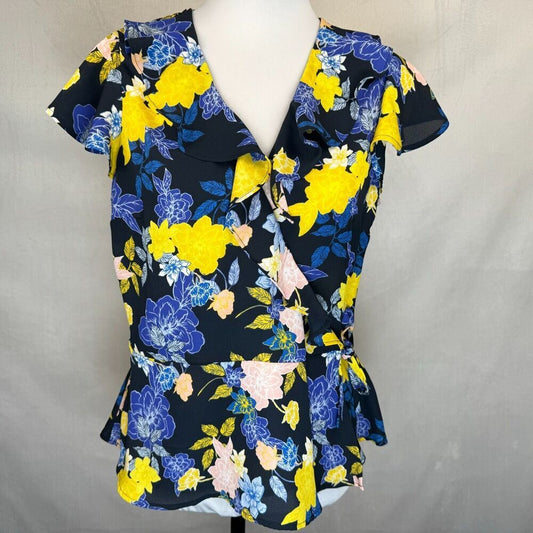 A women's short-sleeve wrap-style blouse with a ruffled V-neck collar, displayed on a white mannequin torso. The blouse features a vibrant floral pattern with bright yellow, blue, and pink flowers on a black background, set against a neutral gray fabric backdrop.