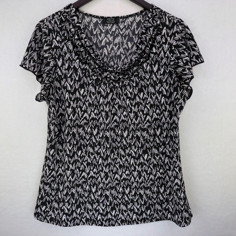 A black and white patterned short-sleeve blouse with a ruffled v-neckline, hanging on a dark wooden hanger against a plain light gray wall.