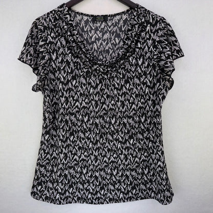 A black and white patterned short-sleeve blouse with a ruffled v-neckline, hanging on a dark wooden hanger against a plain light gray wall.