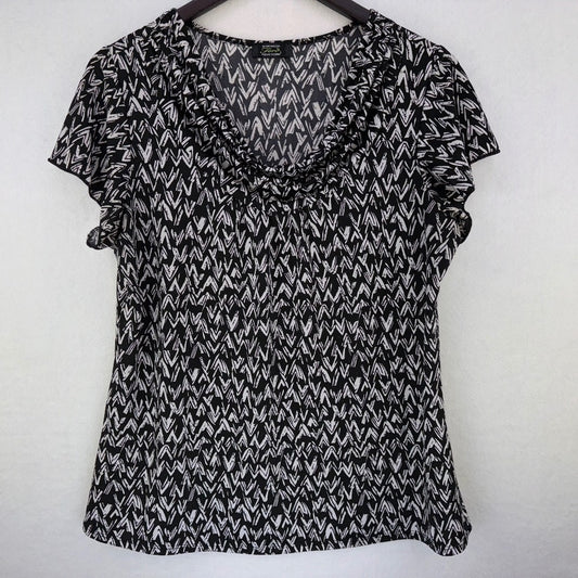A black and white patterned short-sleeve blouse with a ruffled v-neckline, hanging on a dark wooden hanger against a plain light gray wall.