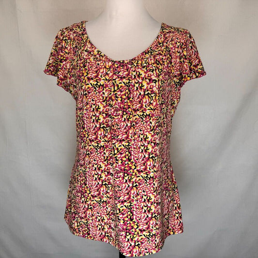 A short-sleeve V-neck blouse with a dense pink, red, yellow, and black floral pattern, displayed on a white mannequin torso against a light gray fabric backdrop.