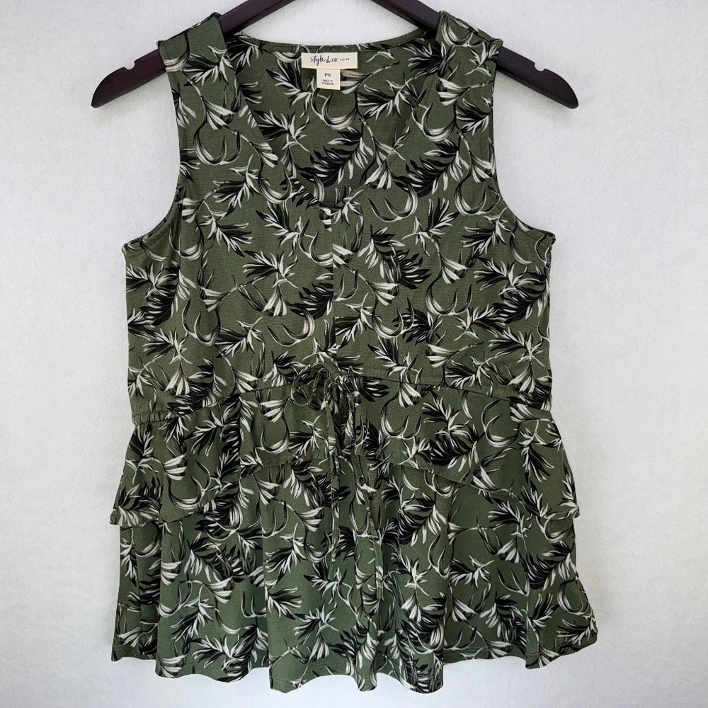An olive green sleeveless ruffled top with a black and white tropical leaf print, hanging on a dark wooden hanger against a light gray textured wall.