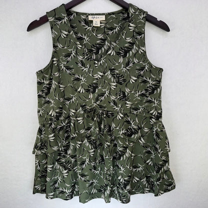 An olive green sleeveless ruffled top with a black and white tropical leaf print, hanging on a dark wooden hanger against a light gray textured wall.