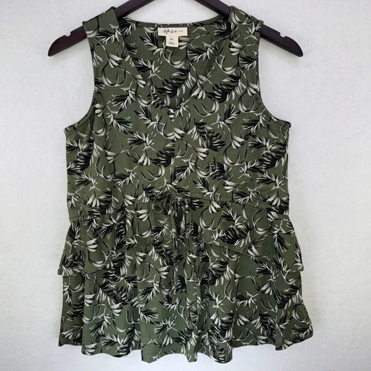 An olive green sleeveless ruffled top with a black and white tropical leaf print, hanging on a dark wooden hanger against a light gray textured wall.