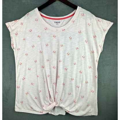 A women's short-sleeve knit top by IZOD, featuring a white heathered base with a small pink and red floral print, a pink inner collar, and a front twist detail, hanging on a dark wooden hanger against a textured gray-green wall.