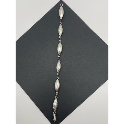 A silver beaded wall hanging or decorative garland with seven elongated textured beads connected by small rings, displayed on a dark gray diamond-shaped card against a light gray background.