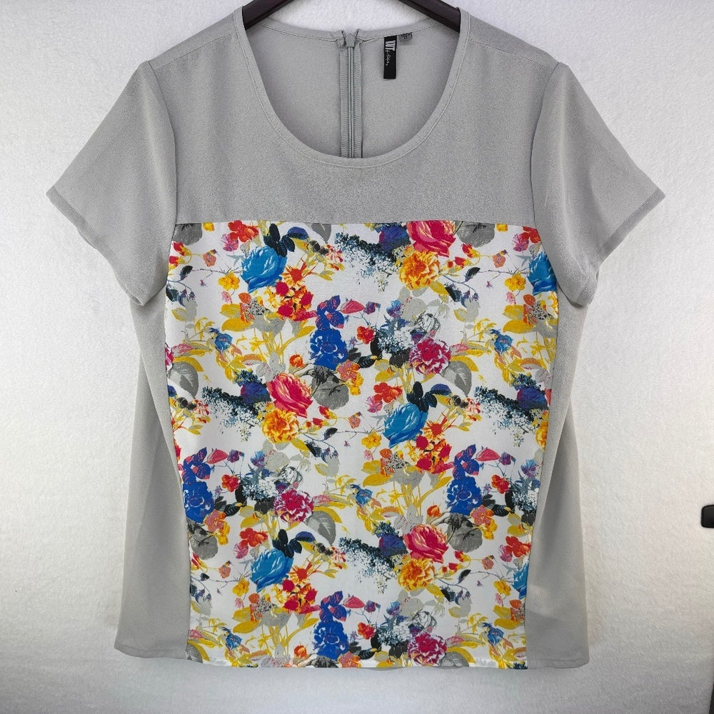 A short-sleeve scoop-neck blouse hanging on a dark hanger against a light textured wall. The top features a solid light gray upper yoke and sleeves, with a large front panel displaying a colorful watercolor floral print on a white background.