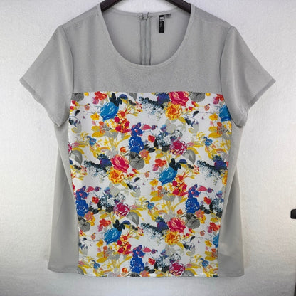 A short-sleeve scoop-neck blouse hanging on a dark hanger against a light textured wall. The top features a solid light gray upper yoke and sleeves, with a large front panel displaying a colorful watercolor floral print on a white background.