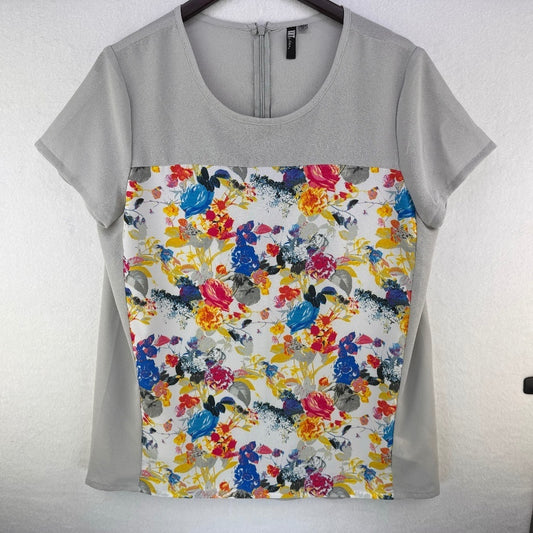 A short-sleeve scoop-neck blouse hanging on a dark hanger against a light textured wall. The top features a solid light gray upper yoke and sleeves, with a large front panel displaying a colorful watercolor floral print on a white background.