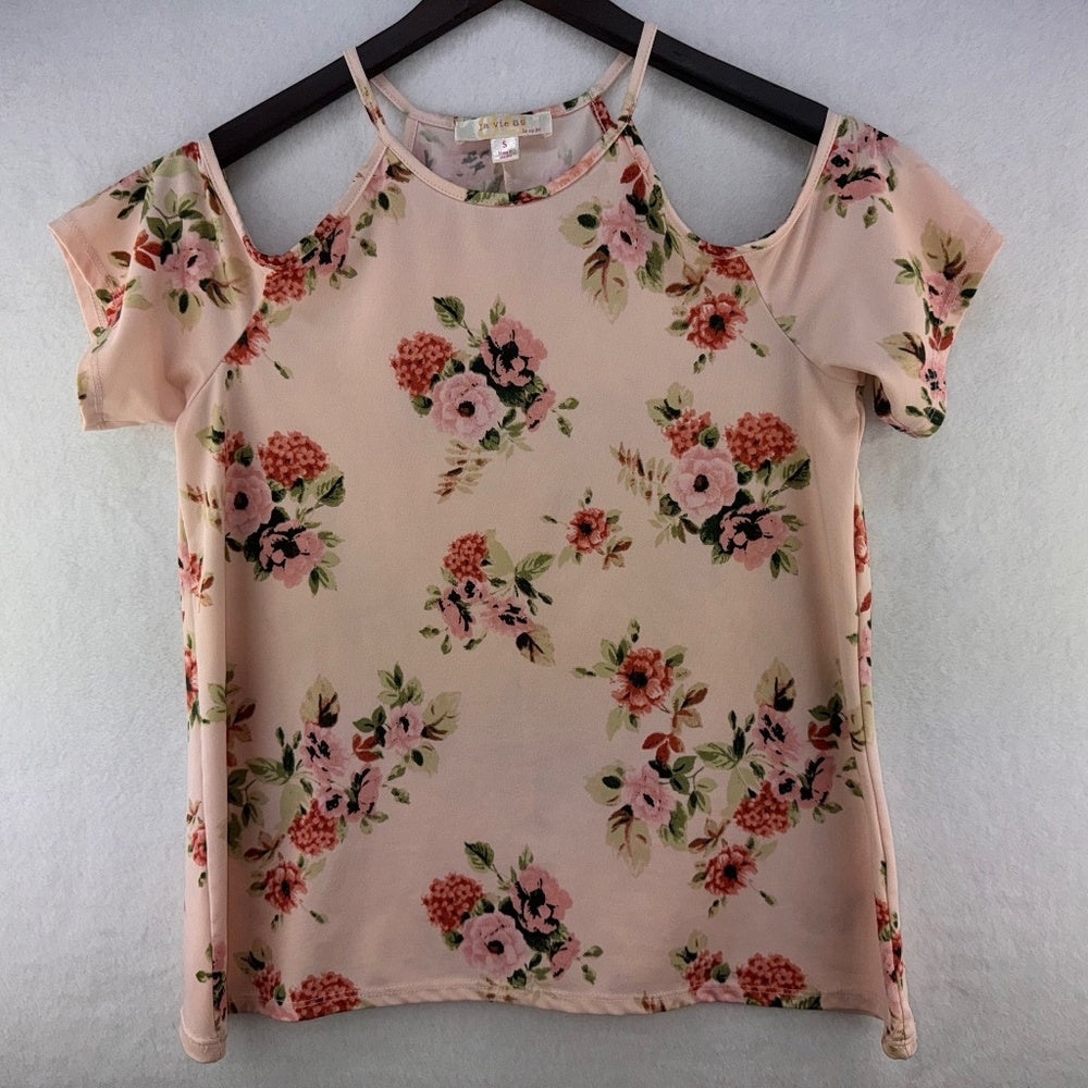 A women's short-sleeve top with a floral print, hanging on a black hanger against a light gray textured wall. The top has a pale pink base with pink, red, and green flowers.