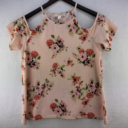 A women's short-sleeve top with a floral print, hanging on a black hanger against a light gray textured wall. The top has a pale pink base with pink, red, and green flowers.