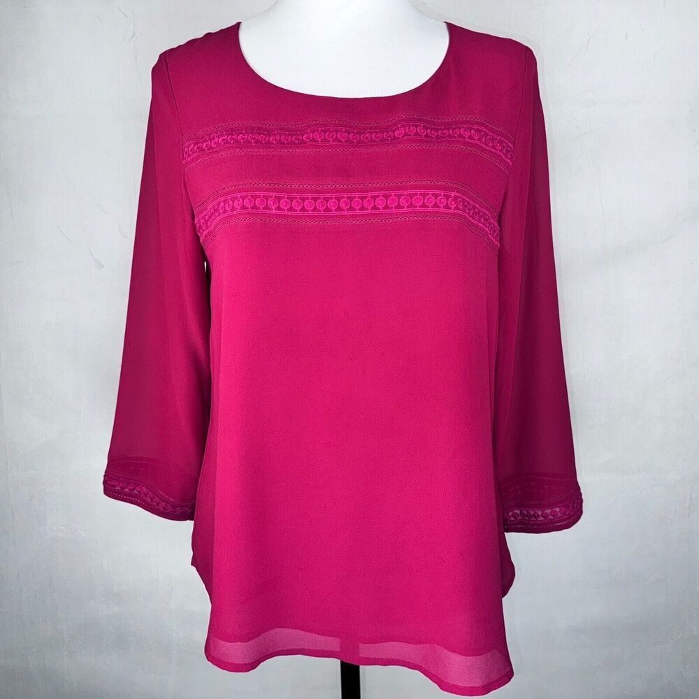 A bright magenta pink long-sleeve blouse with decorative embroidered horizontal bands across the chest, displayed on a white mannequin torso against a light gray background.
