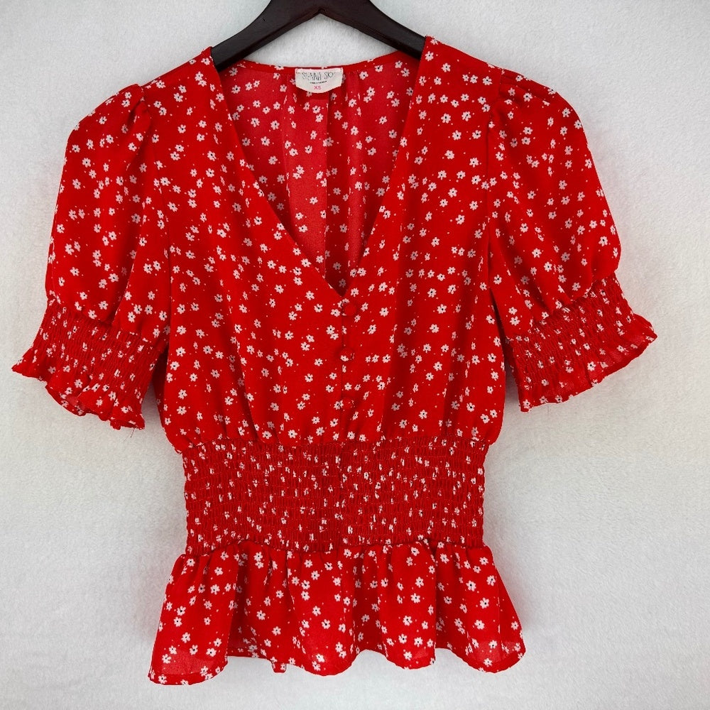 A red short-sleeve blouse with a white ditsy floral print, featuring puff sleeves, a smocked waist, and a ruffled hem, hanging on a black hanger against a textured white wall.