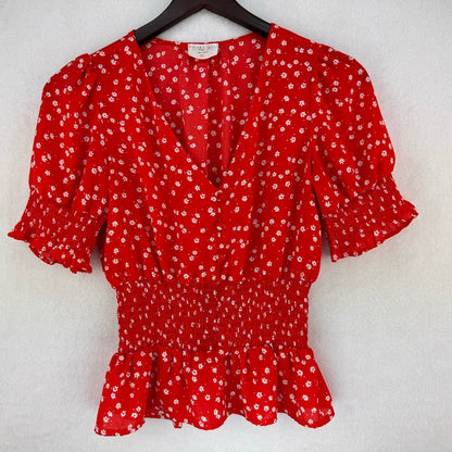 A red short-sleeve blouse with a white ditsy floral print, featuring puff sleeves, a smocked waist, and a ruffled hem, hanging on a black hanger against a textured white wall.