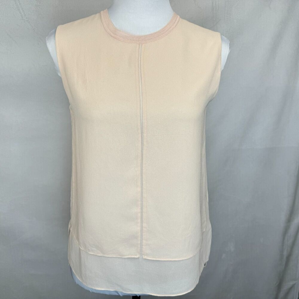 A sleeveless cream-colored top with a round neckline and a central seam, displayed on a white mannequin torso against a gray fabric backdrop.