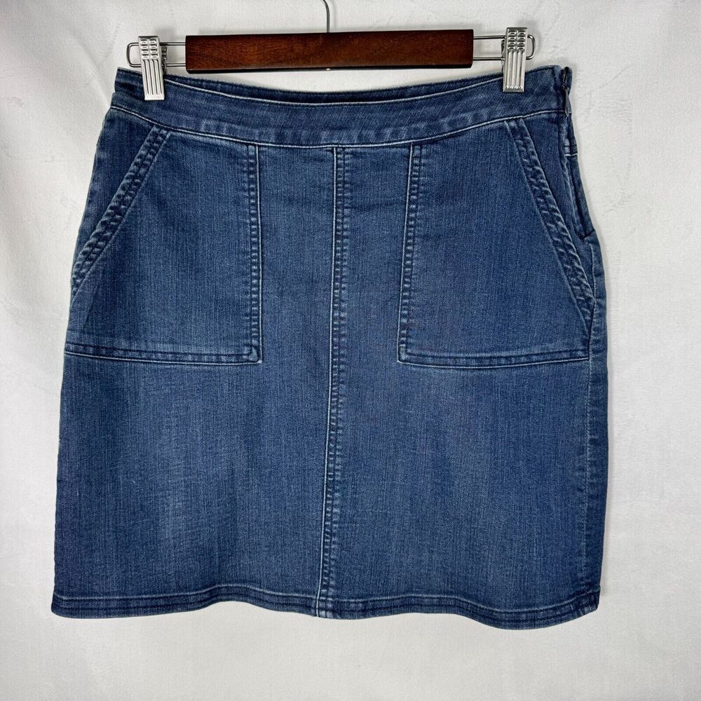 A blue denim mini skirt with front patch pockets, hanging on a wooden hanger against a white fabric background.