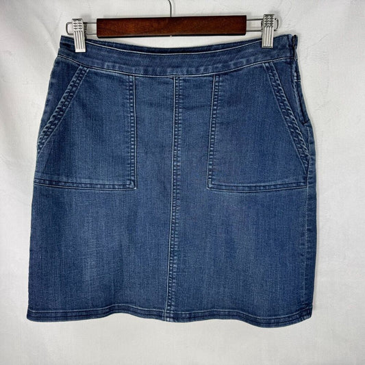 A blue denim mini skirt with front patch pockets, hanging on a wooden hanger against a white fabric background.