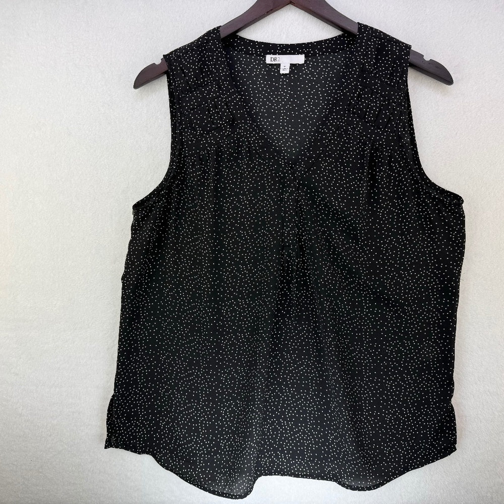 A sleeveless black V-neck blouse with a small white speckled pattern, hanging on a dark wooden hanger against a textured off-white wall.