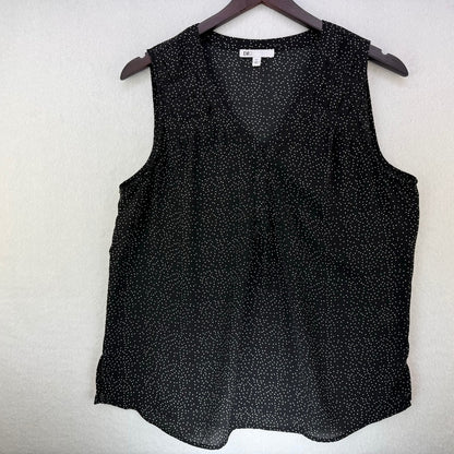 A sleeveless black V-neck blouse with a small white speckled pattern, hanging on a dark wooden hanger against a textured off-white wall.