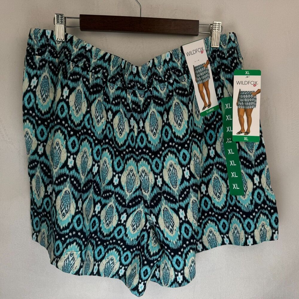 A pair of women's shorts with a teal, black, and white ikat pattern, hanging on a wooden hanger with retail tags attached. The brand 'WildFashion' is visible on the tags.