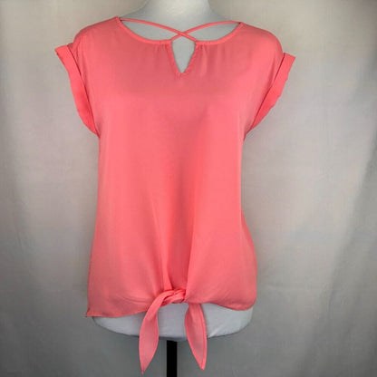 A coral pink short-sleeve blouse with a keyhole back and front tie detail, displayed on a white mannequin torso against a light gray fabric backdrop.