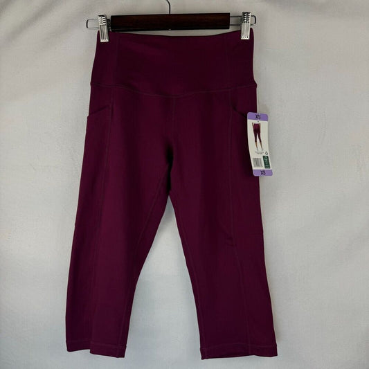 A pair of women's high-waisted capri leggings in deep burgundy, hanging on a black hanger against a light gray fabric backdrop. A product tag is attached to the right side.
