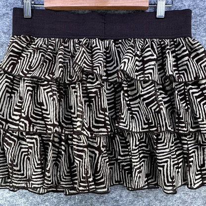 A tiered ruffle mini skirt with a black and beige geometric maze-like pattern, featuring a solid black waistband, hanging on a wooden hanger against a gray textured background.