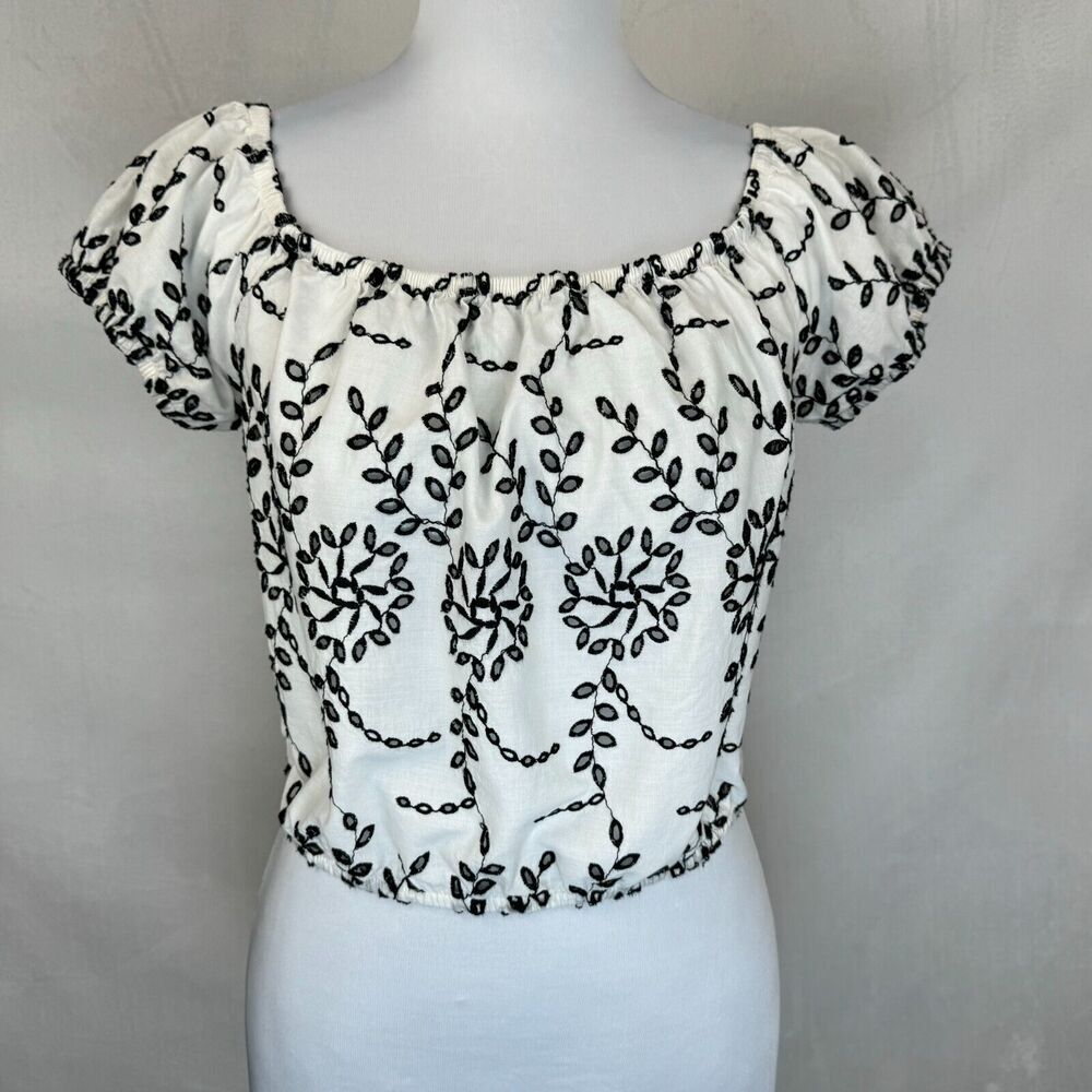A white short-sleeve blouse with black floral embroidery, displayed on a white mannequin torso against a light gray background.