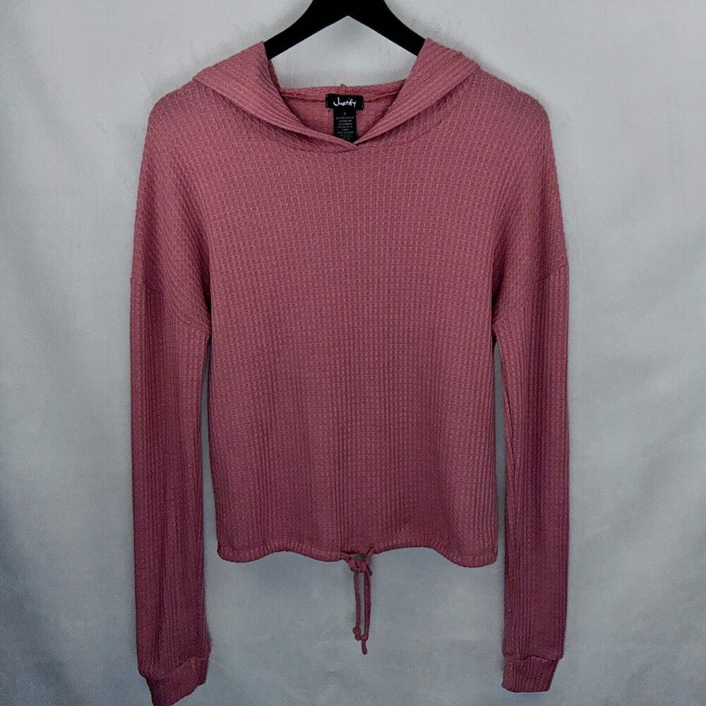A dusty rose pink ribbed knit cropped hoodie sweater with a drawstring hem, hanging on a black hanger against a light gray fabric backdrop.