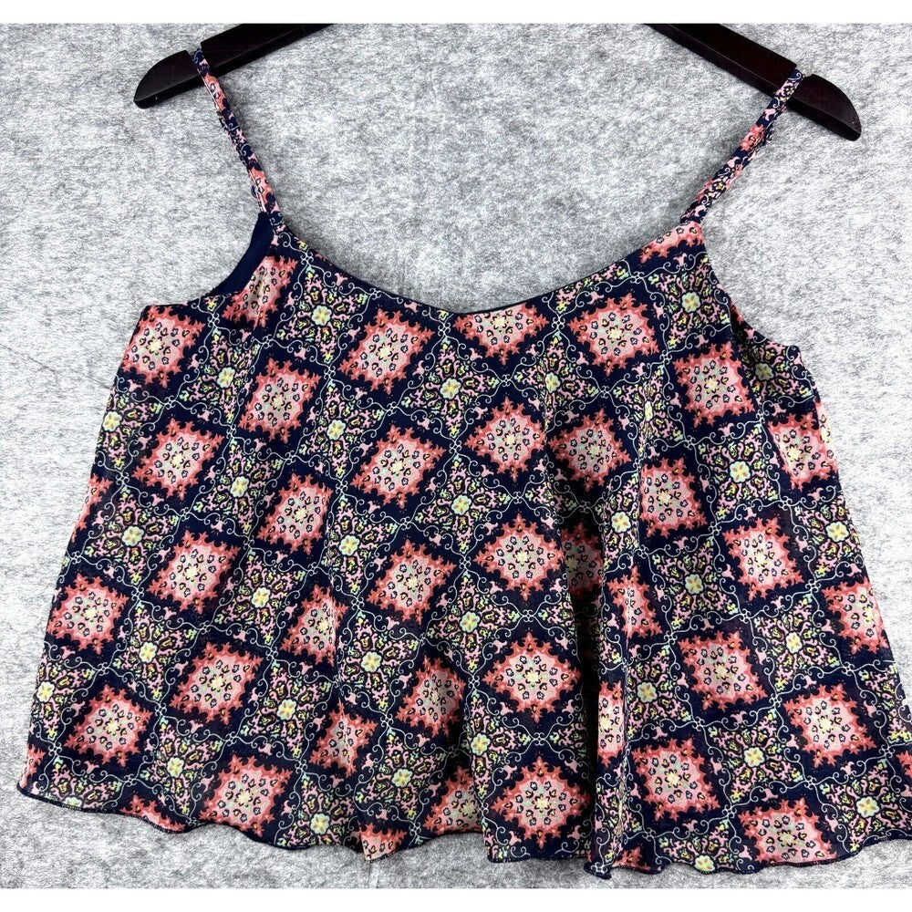 A sleeveless spaghetti-strap top with a colorful geometric and floral pattern, hanging on a dark wooden hanger against a textured gray background.