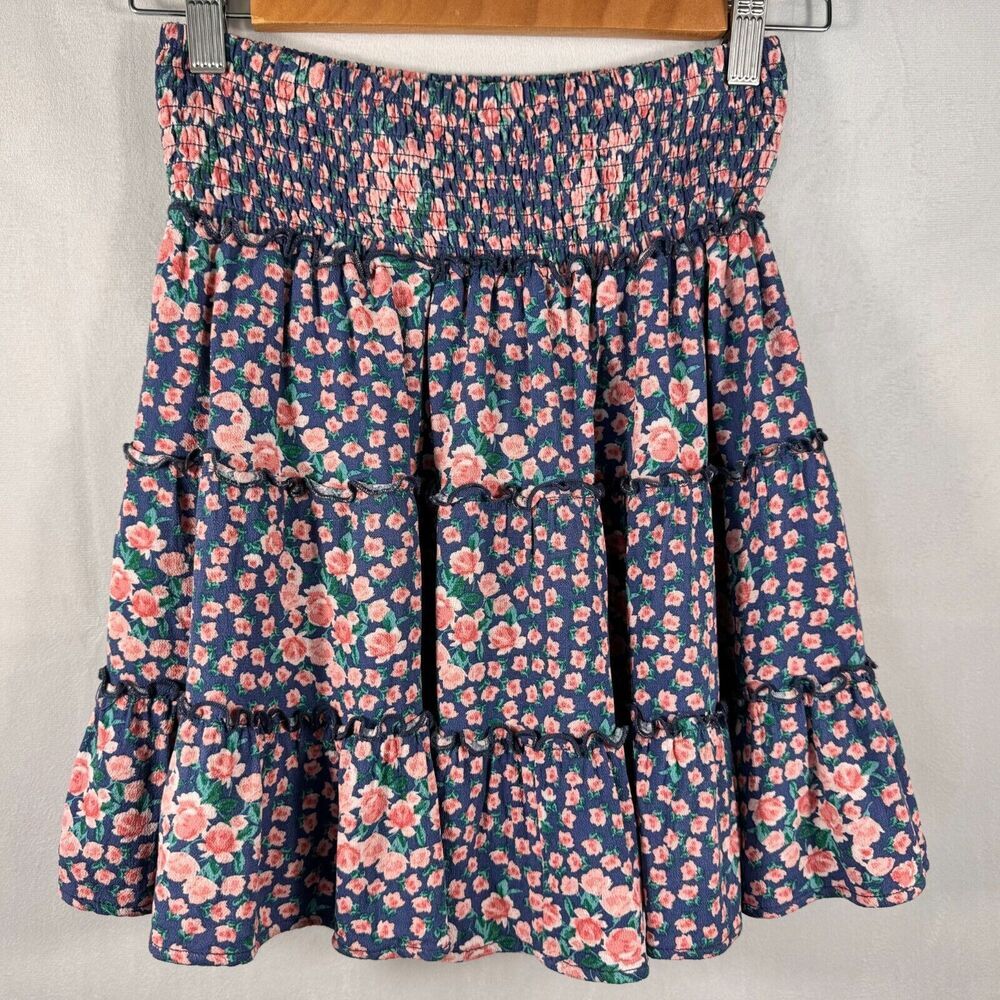 A tiered floral skirt with a smocked elastic waistband, featuring a navy blue base with pink and green floral patterns, hanging on a wooden hanger against a light gray fabric backdrop.