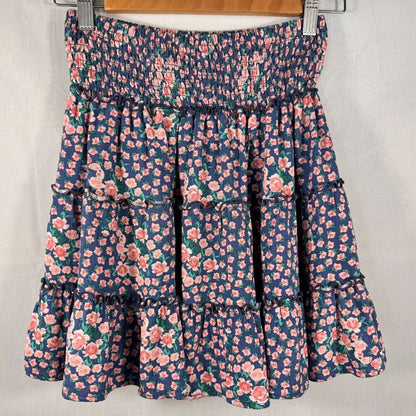 A tiered floral skirt with a smocked elastic waistband, featuring a navy blue base with pink and green floral patterns, hanging on a wooden hanger against a light gray fabric backdrop.