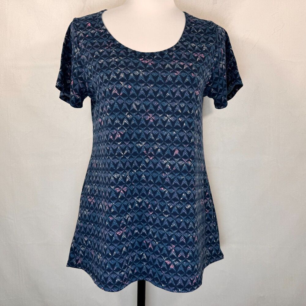 A short-sleeve scoop-neck tunic top in navy blue with a repeating geometric pattern of pink, purple, and white shapes, displayed on a white mannequin torso against a white fabric backdrop.
