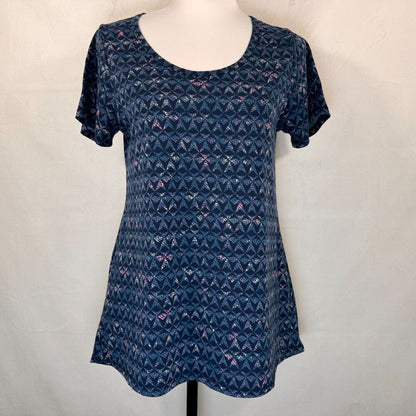 A short-sleeve scoop-neck tunic top in navy blue with a repeating geometric pattern of pink, purple, and white shapes, displayed on a white mannequin torso against a white fabric backdrop.