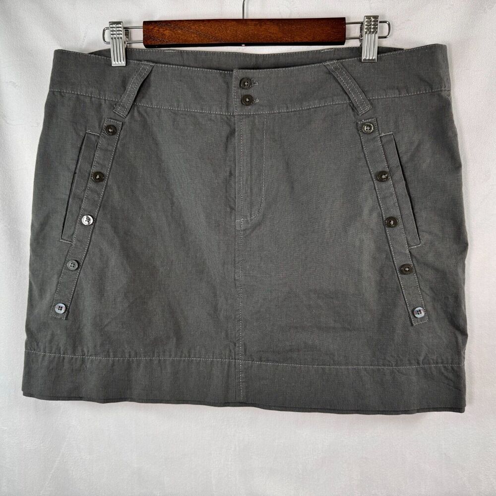 A gray mini skirt with buttoned side pockets, hanging on a wooden hanger against a white fabric background.