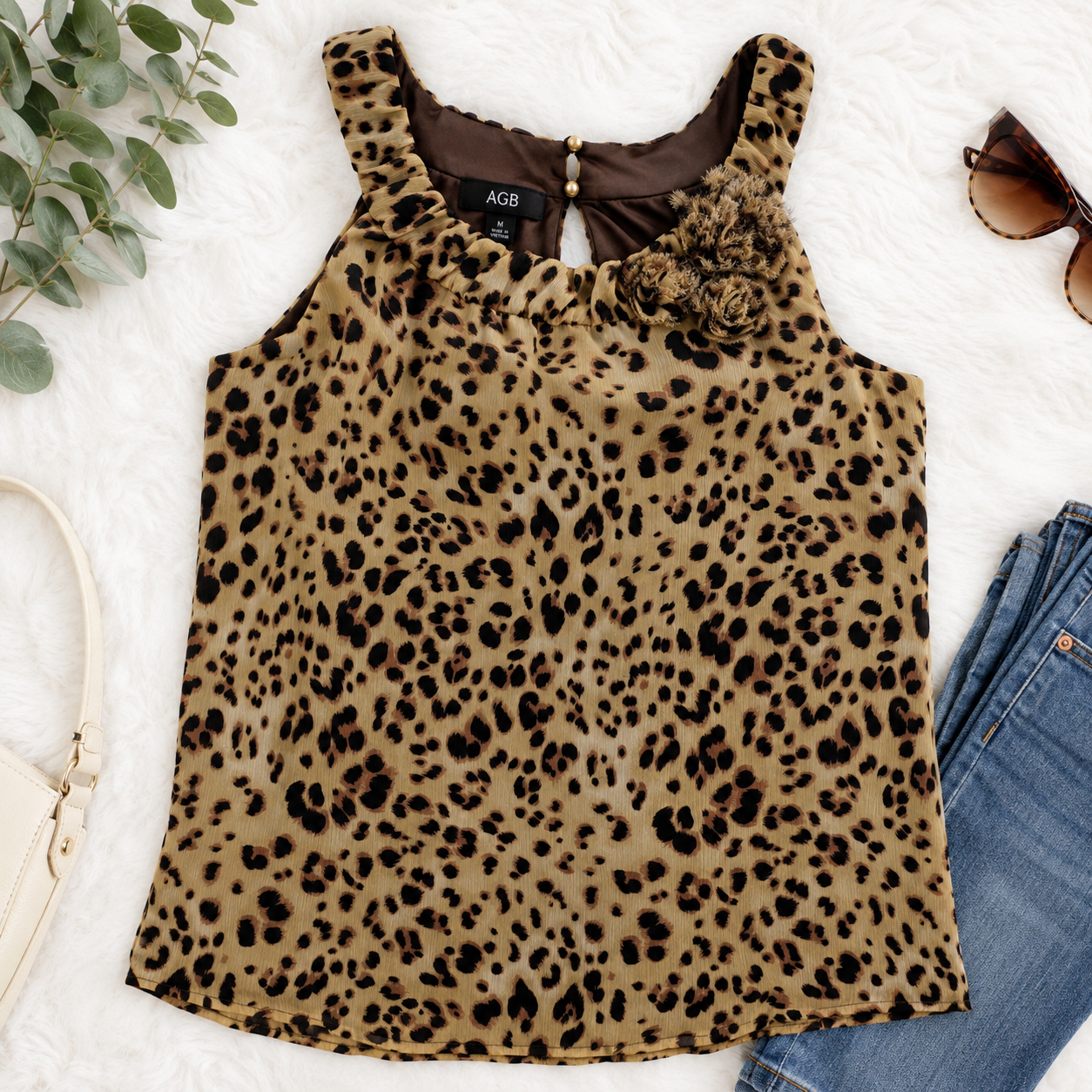A sleeveless leopard-print top with a ruffled neckline and a 3D floral embellishment on the chest, styled in a flat lay with jeans, sunglasses, a handbag, and green leaves on a white textured background. The brand label reads ACGB.