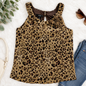 A sleeveless leopard-print top with a ruffled neckline and a 3D floral embellishment on the chest, styled in a flat lay with jeans, sunglasses, a handbag, and green leaves on a white textured background. The brand label reads ACGB.