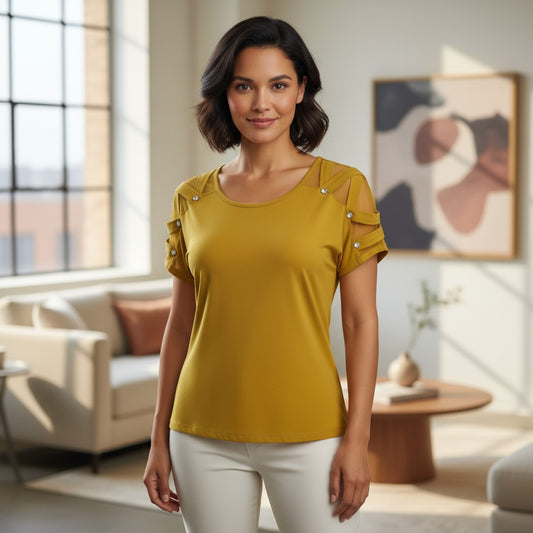 A woman wearing a mustard yellow short-sleeve top with decorative silver stud details on the shoulders and sleeves, paired with white pants, standing in a modern, sunlit living room.