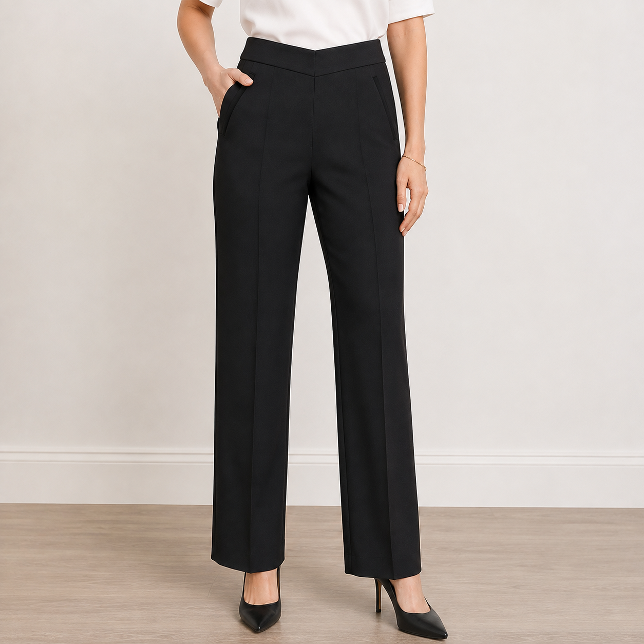 A model wearing black wide-leg trousers with a white top and black heels, standing against a neutral beige wall and light wood floor.
