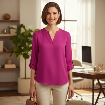 A woman stands in a bright home office wearing a bright magenta long-sleeve blouse with a split V-neck and rolled sleeves, paired with light beige pants.