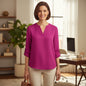 A woman stands in a bright home office wearing a bright magenta long-sleeve blouse with a split V-neck and rolled sleeves, paired with light beige pants.