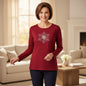 A smiling woman stands in a bright living room wearing a long-sleeve crewneck top in deep red with a white and gold embroidered snowflake design on the chest, paired with dark navy pants.