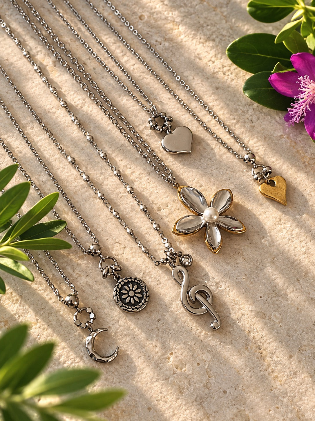 A set of silver-toned and gold-toned delicate necklaces with various charms, including a flower, treble clef, crescent moon, and hearts, arranged on a textured beige stone surface with green leaves and a purple flower.