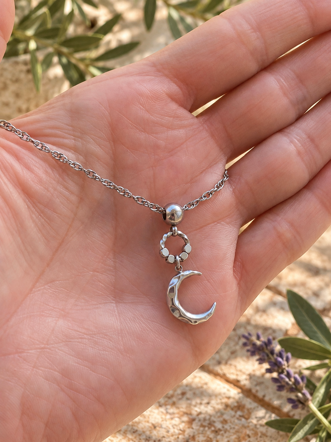 A sterling silver necklace with a crescent moon and sun pendant is held in a person's hand, set against a blurred background of olive trees and sand.