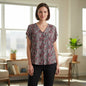 A woman stands in a bright, modern room wearing a short-sleeve V-neck blouse with a dense red, pink, and white floral print on a dark background, paired with black pants.