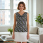 A woman stands in a bright living room wearing a sleeveless V-neck top with a black, white, and olive green floral pattern, paired with a white skirt.