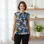 A woman wearing a sleeveless black blouse with a blue, white, and light purple floral print, paired with black pants, standing in a bright, modern room with shelves and a plant.